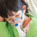 Child in lime green sweater with an nebulizer mask on while lying dow with eyes closed.